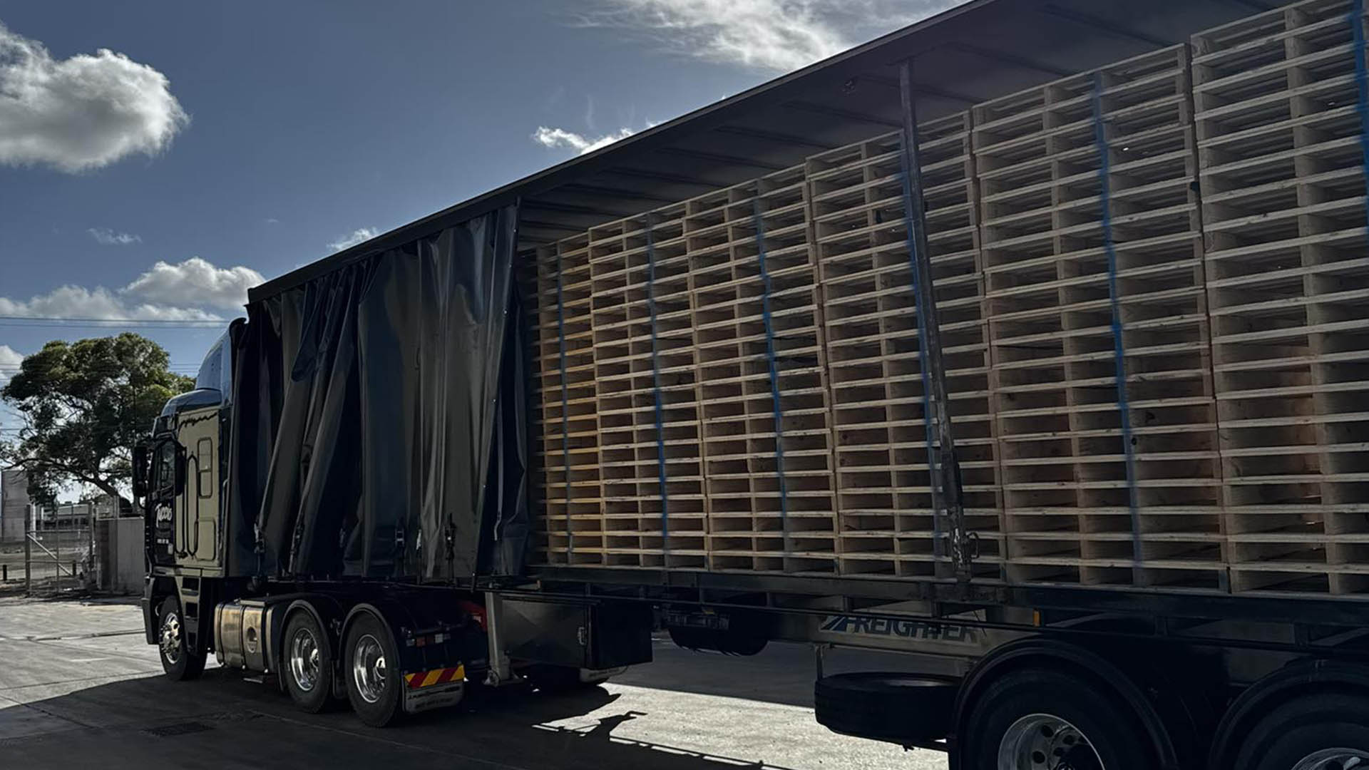 1-home-bulk Wide view of a curtain-sider semi-trailer with curtains pulled back, loaded with timber pallets