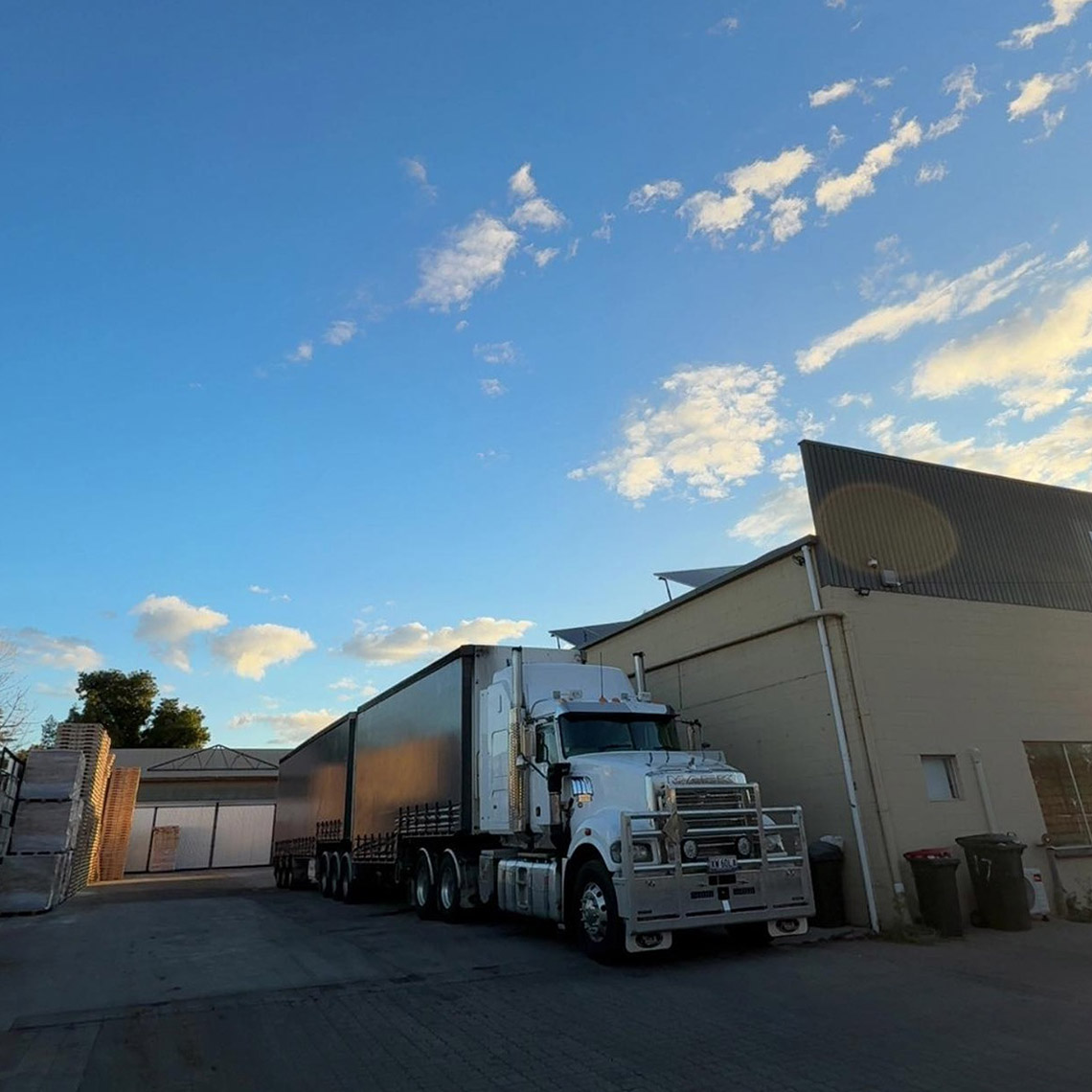 White prime mover with two curtain-sider trailers parked beside a shed