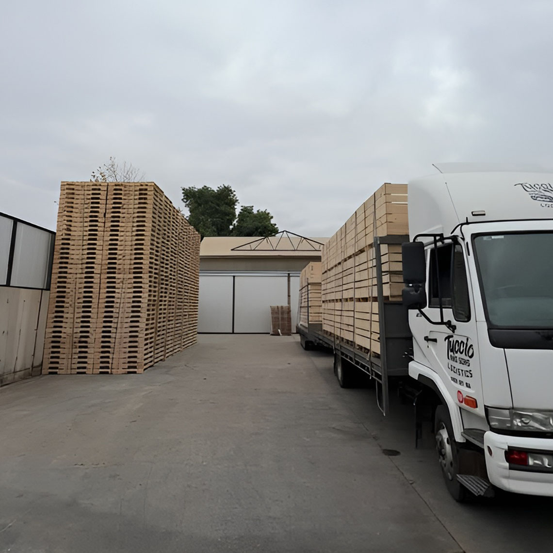 White rigid truck loaded with pallets beside tall pallet stacks in a concrete yard