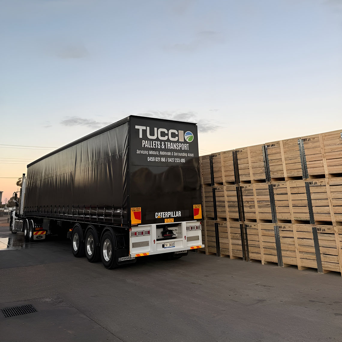 Black curtain-sider semi-trailer parked beside stacked timber bins at dusk