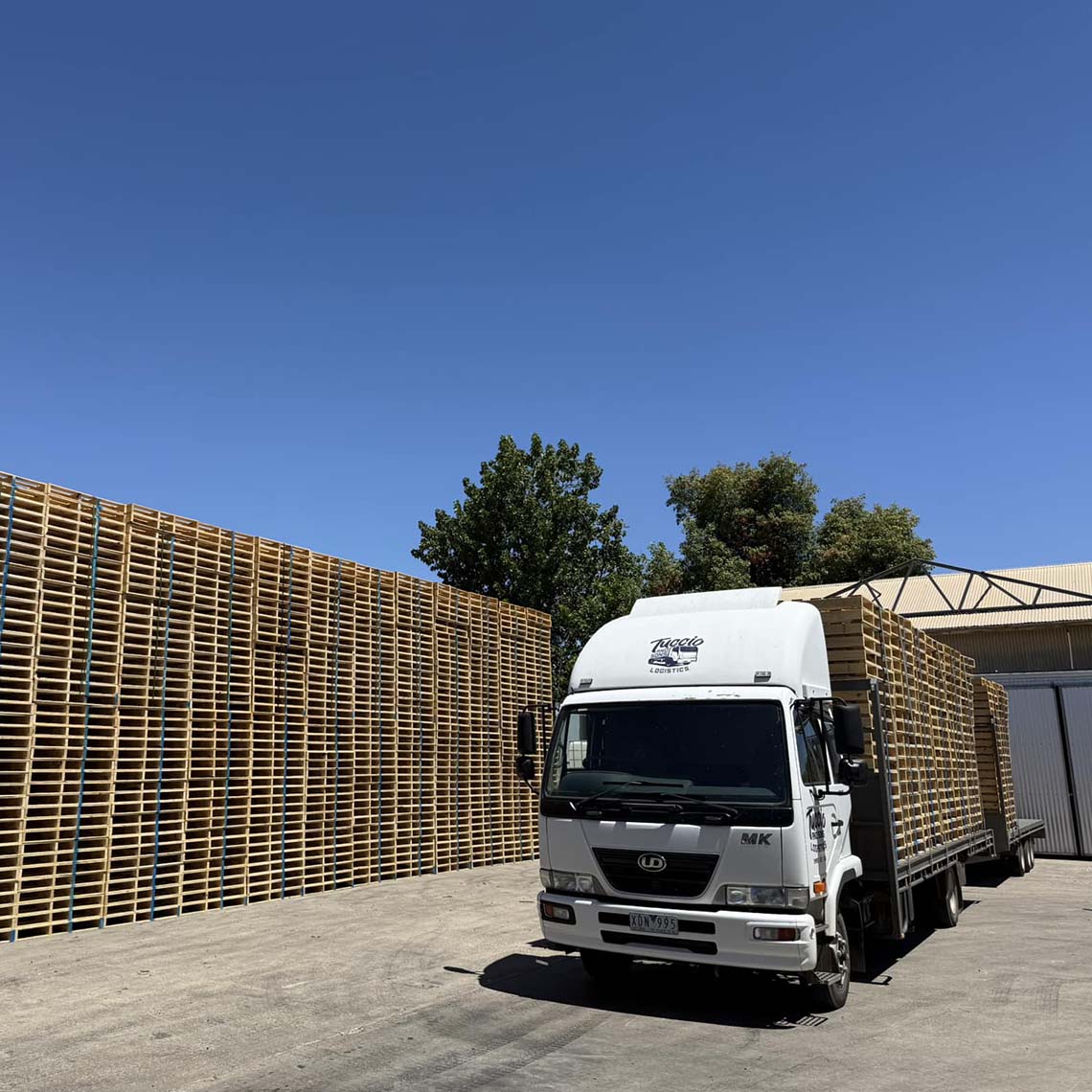 White rigid truck loaded with pallets in front of a long wall of stacked pallets
